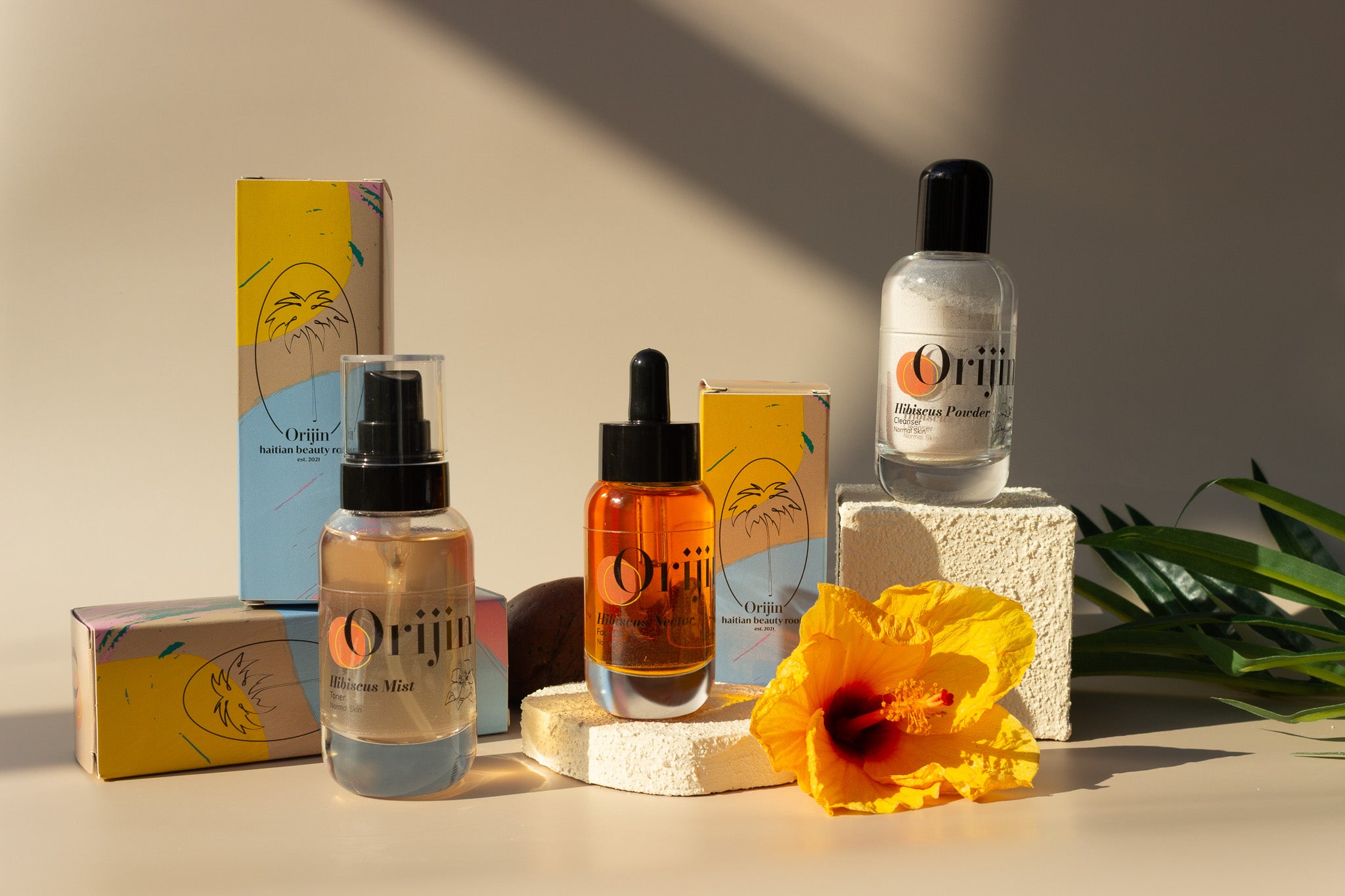 A collection of skincare products including a powder, mist, and oil, with packaging that features hibiscus imagery and a sunset hue. A fresh hibiscus flower and leaves are also present in the image.